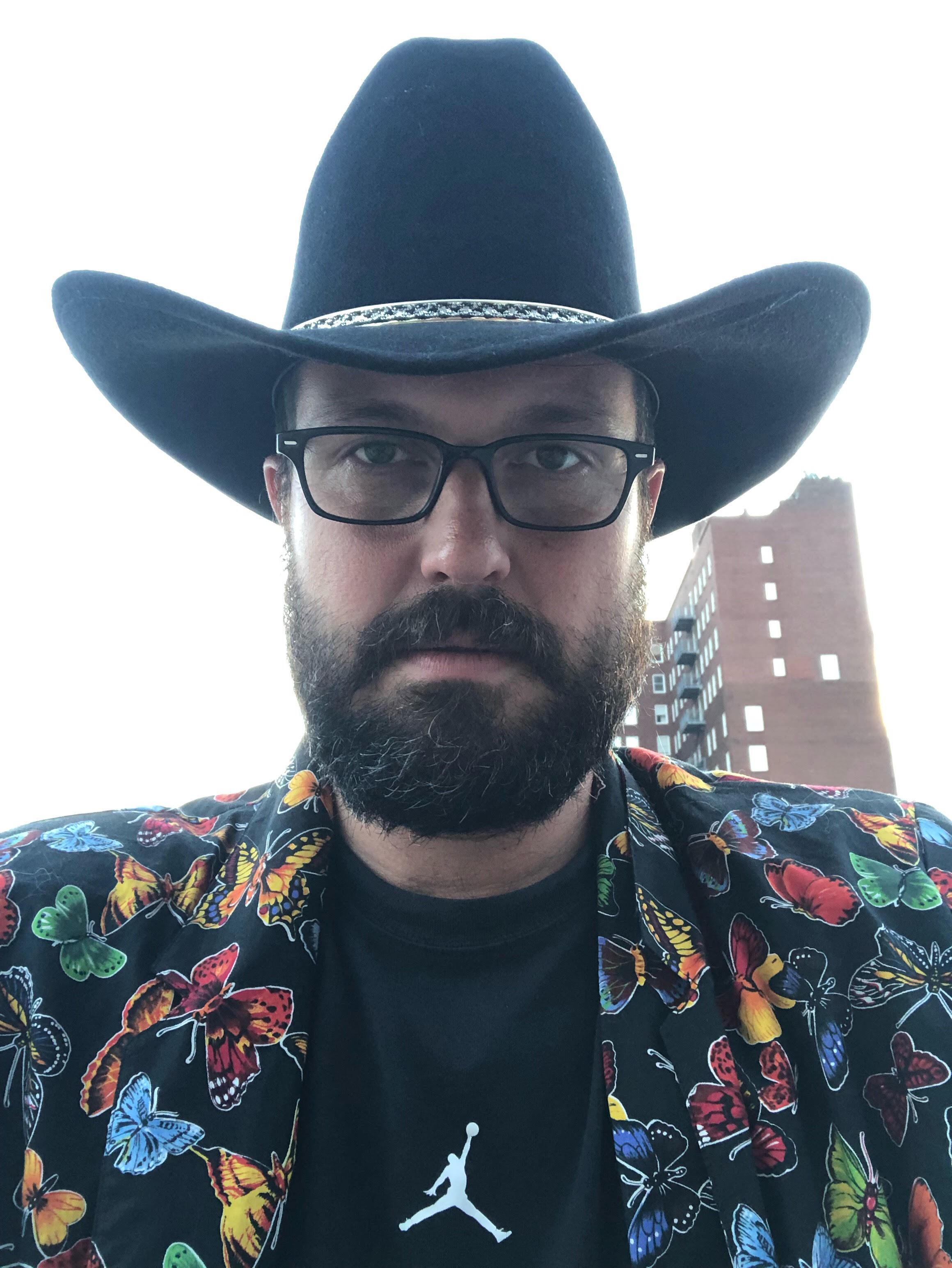 David wears a wide brimmed hat, has black framed glasses, and sports a full beard. He is wearing button up with a colorful assortment of butterflies on top of a tshirt with the air jordan logo.