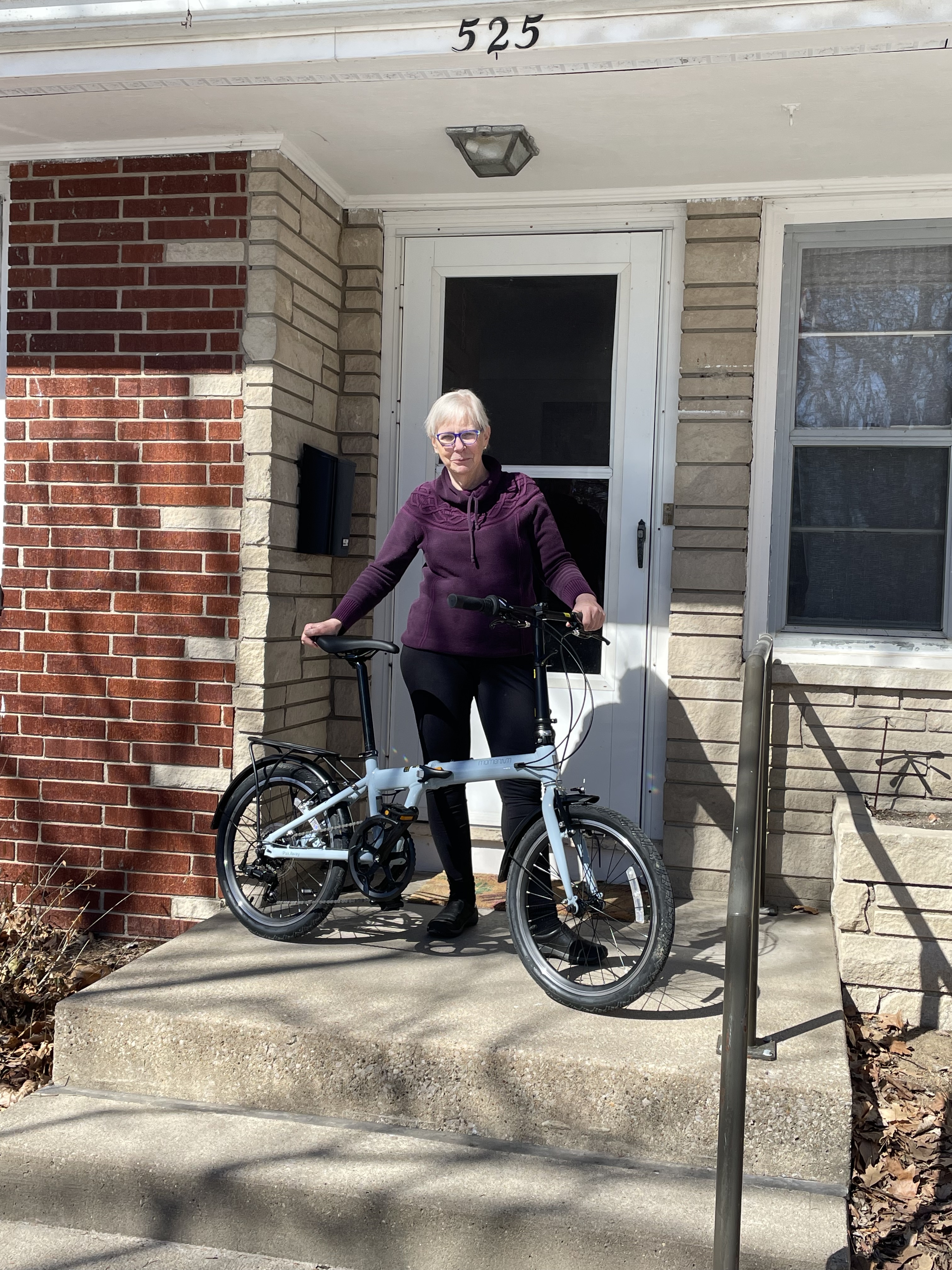 Margee stands on the front steps of a house. She is wearing a purple fleece and black athletic pants. She holds onto a powder blue foldable bike.